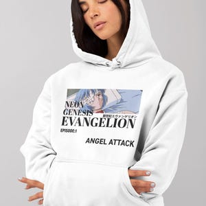 May include: White hoodie featuring a graphic from the anime series Neon Genesis Evangelion. The design includes the show's title, Japanese text, and the phrase "ANGEL ATTACK." The hoodie has a drawstring hood and a front pocket.