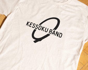 May include: A cream-colored short-sleeve t-shirt featuring a black graphic print on the chest. The design consists of the text KESSOKU BAND written in a bold, sans-serif font, partially encircled by a thick, black, hand-drawn style oval shape.