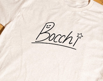 May include: A cream-colored short-sleeve t-shirt featuring a black graphic print on the chest. The design displays the word Bocchi in a handwritten script font, with a small smiling face on the letter B and a star symbol at the end.