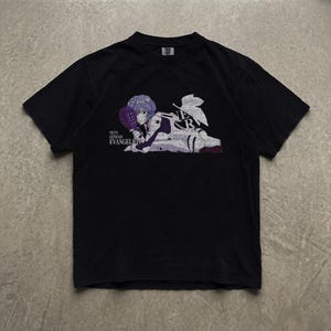 May include: A black short-sleeved t-shirt featuring a graphic print of a character with short blue hair lying down while holding a purple paddle. The text Neon Genesis Evangelion and NERV are printed in white near the character illustration.