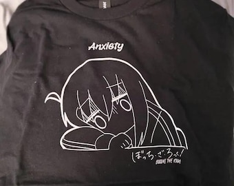 May include: A black t-shirt featuring a white line art graphic of a character resting their head on their arms. Above the graphic is the word "Anxiety" in white text. Below the graphic is the text "ぼっち・ざ・ろっく!" and "BOCCHI THE ROCK!" in white.