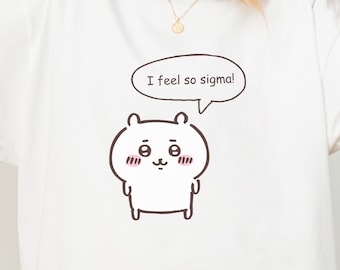 Puede incluir: Una camiseta blanca con una ilustración de líneas marrones de una pequeña criatura redonda de dibujos animados con mejillas rosadas y un bocadillo con el texto I feel so sigma! El diseño está centrado en el pecho de la prenda.