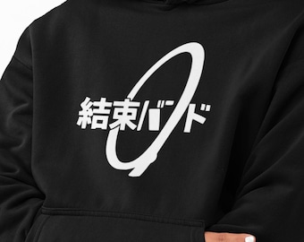 May include: A black hooded sweatshirt featuring a white graphic print on the chest. The design consists of a large white tilted oval shape overlapping Japanese text that reads 結束バンド. The hoodie includes a front kangaroo pocket and a drawstring hood.