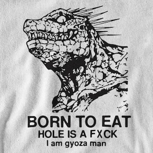 May include: A white t-shirt featuring a black graphic print of a spiky, reptilian creature. Below the creature, the text reads: BORN TO EAT, HOLE IS A FXCK, I am gyoza man. The fabric has a visible texture and the print is high contrast.