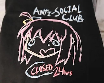 May include: A black t-shirt featuring a graphic print of a stylized anime-style character with pink hair and a distressed expression. The text "ANTI-SOCIAL CLUB" appears above the character, and the text "CLOSED 24hrs" is printed below in a sketch style.