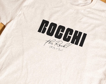 May include: A cream-colored short-sleeve t-shirt featuring the black text "BOCCHI" in a bold, sans-serif font. Below this, the text "the Rock!" is written in a cursive script, followed by smaller Japanese characters in a handwritten style.