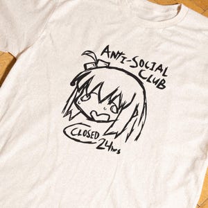 May include: A cream-colored short-sleeve t-shirt featuring a black line-art graphic of an anime-style character head. The text reads ANTI-SOCIAL CLUB above the character and CLOSED 24hrs inside an oval shape below the character's chin.