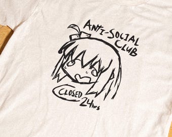 May include: A cream-colored short-sleeve t-shirt featuring a black line-art graphic of an anime-style character head. The text reads ANTI-SOCIAL CLUB above the character and CLOSED 24hrs inside an oval shape below the character's chin.