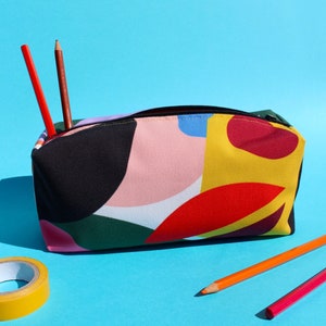 Puede incluir: Un estuche para l&aacute;pices colorido con un patr&oacute;n geom&eacute;trico en tonos de rosa, amarillo, rojo, verde y negro. El estuche est&aacute; hecho de lona y tiene una cremallera. Dos l&aacute;pices sobresalen del estuche.