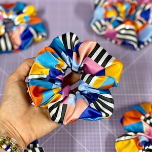 Könnte beinhalten: Ein farbenfroher Haargummi mit geometrischem Muster mit schwarzen und weißen Streifen, blau, rosa, orange und gelb.