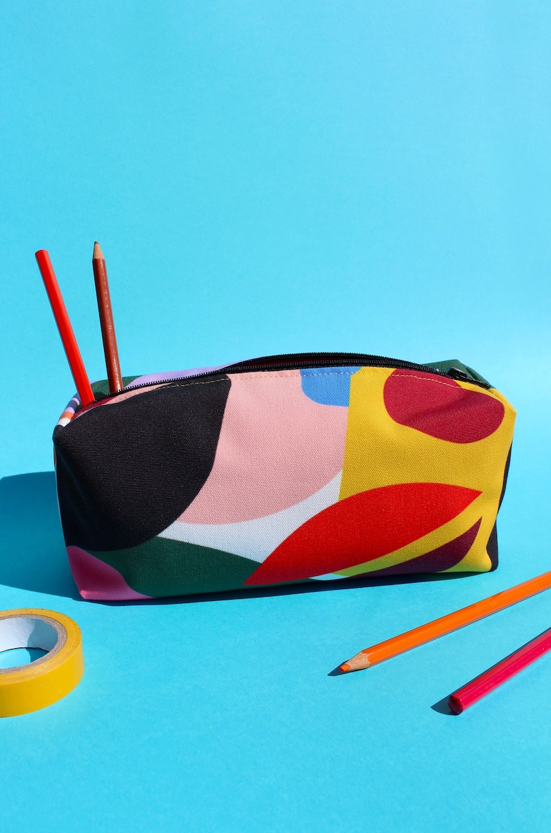 Puede incluir: Un estuche para l&aacute;pices colorido con un patr&oacute;n geom&eacute;trico en tonos de rosa, amarillo, rojo, verde y negro. El estuche est&aacute; hecho de lona y tiene una cremallera. Dos l&aacute;pices sobresalen del estuche.
