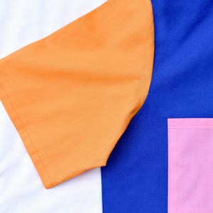 以下が含まれることがあります： 青、オレンジ、ピンクのカラーブロックデザインの半袖シャツ。シャツの青い部分にピンクのポケットが付いている。