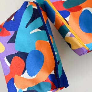 Può includere: Due astucci rettangolari con cerniera caratterizzati da un vivace motivo astratto di forme organiche nei toni del blu, arancione, rosso, viola e giallo. Uno ha una cerniera blu scuro, mentre l'altro presenta una chiusura giallo brillante.