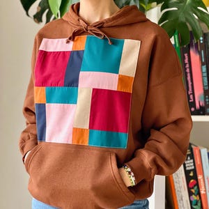 Peut inclure: Un sweat-shirt marron avec un grand motif patchwork sur le devant. Le patchwork est composé de carrés de différentes couleurs, dont le rouge, le rose, le bleu, le vert, l'orange et le blanc.