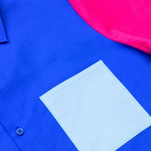 以下が含まれることがあります： ボタンダウンの青いシャツで、薄い青色のポケットとピンク色の袖が付いています。