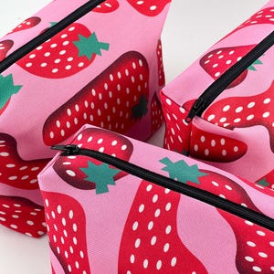 Peut inclure: Trois trousses rectangulaires en tissu avec un motif répétitif de fraises rouges à graines blanches et feuilles vertes sur fond rose clair. Chaque trousse est dotée d'une fermeture éclair noire sur le bord supérieur pour le rangement.