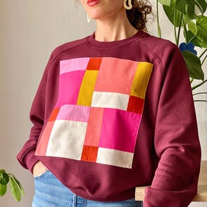 May include: A maroon sweatshirt with a large patchwork design on the front. The patchwork is made up of squares of pink, red, yellow, and white fabric.
