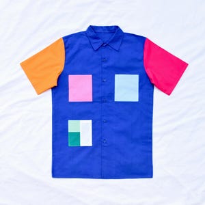 Peut inclure: Une chemise boutonnée à manches courtes de couleur bleue avec des carrés orange, rose et bleu clair sur le devant. La chemise a un col et cinq boutons sur le devant.