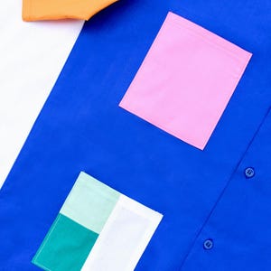 以下が含まれることがあります： 3つのボタンと2つのパッチポケットが付いた青いボタンダウンシャツ。1つのポケットはピンクで、もう1つは緑、白、薄い緑のパッチワークです。