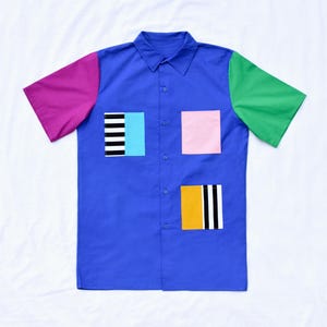 May include: A blue short-sleeved button-down shirt with colourful geometric patches. The patches are pink, green, yellow, black and white.