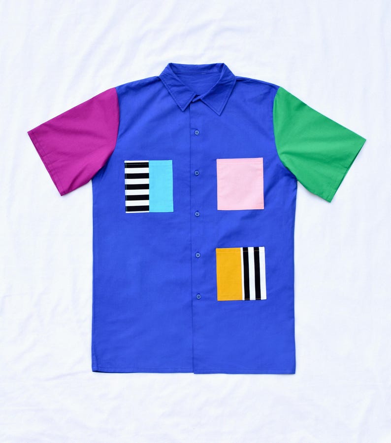 May include: A blue short-sleeved button-down shirt with colourful geometric patches. The patches are pink, green, yellow, black and white.