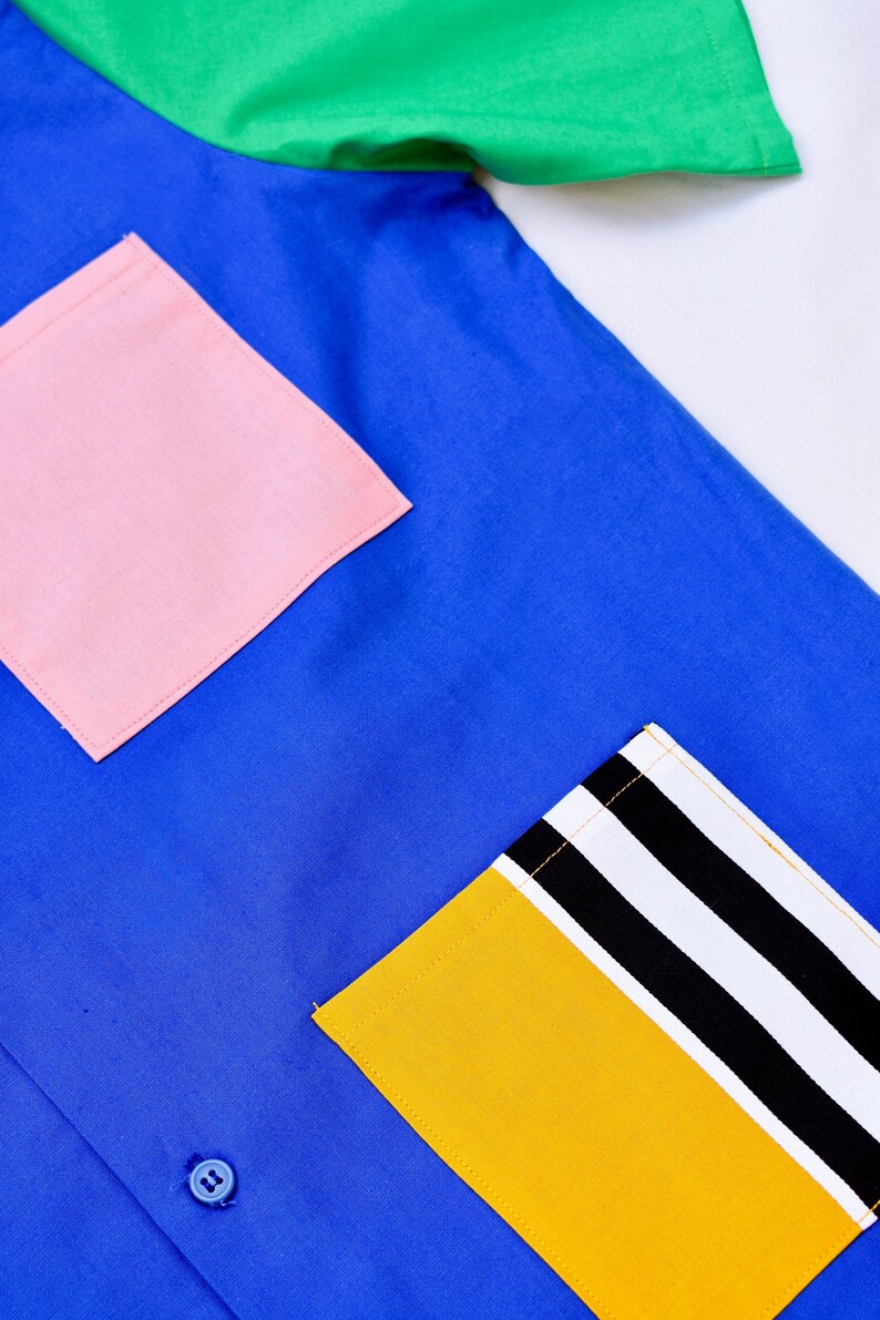 May include: A blue shirt with a pink pocket and a yellow pocket with black and white stripes. The shirt has a single button.