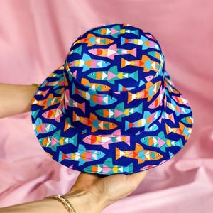 Op de afbeelding: Een blauwe bucket hat met een kleurrijk vispatroon. De vissen zijn gemaakt van geometrische vormen in tinten roze, oranje, geel, groen en wit.