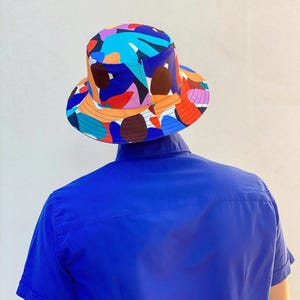 Peut inclure: Un chapeau bob orné d'un motif abstrait multicolore vif avec des formes dans des tons de bleu, orange, rouge, violet et marron. Le chapeau présente un sommet plat et un bord incliné vers le bas, porté au-dessus d'une chemise bleue unie.
