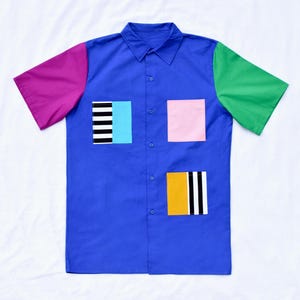 May include: A blue short-sleeved button-down shirt with a pink, green, and black and white striped patchwork design.