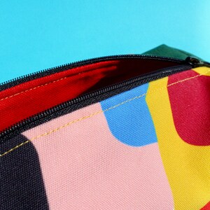 Puede incluir: Una bolsa con cremallera colorida con un patr&oacute;n geom&eacute;trico en tonos de rojo, amarillo, azul, rosa y negro. La bolsa est&aacute; abierta, revelando un forro rojo.