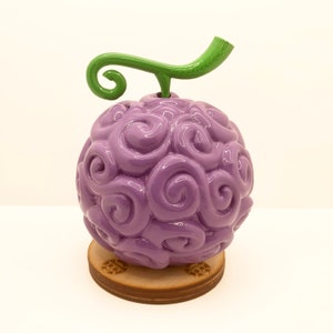 Puede incluir: Una figura de color morado y verde del "Fruta Gomu Gomu" de la serie de anime One Piece. La fruta está sobre una base de madera.