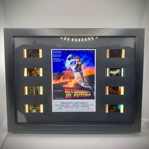 Può includere: Un poster cinematografico incorniciato di "Ritorno al Futuro" con strisce di pellicola. Il poster presenta un uomo in abito futuristico su sfondo blu e arancione. La cornice è nera.