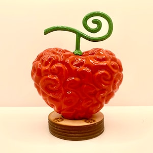 Peut inclure: Une figurine rouge en forme de cœur avec un tourbillon vert sur le dessus, ressemblant à un fruit. La figurine est sur une base en bois.