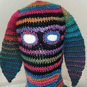 May include: A colorful crocheted mask with two long ear flaps. The mask has a striped pattern in shades of pink, black, blue, green, and orange. The eyes are cut out and lined with white fabric.