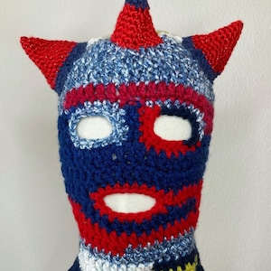 May include: A handmade crochet balaclava in a superhero mask design. The mask features a blue, red, and white color scheme with pointed red horns and eye openings. The balaclava is designed to cover the head and neck.