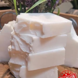 May include: A stack of white, rectangular soap bars with a rough, textured appearance. The bars are stacked on a wooden surface, with a blurred background of purple flowers and greenery. A red "Best Seller" sticker is in the bottom right corner.