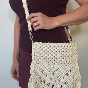 May include: Cream-colored macrame crossbody bag with a woven design and fringe detail. The bag has a braided strap and silver-tone hardware. The bag is a stylish accessory for carrying essentials.