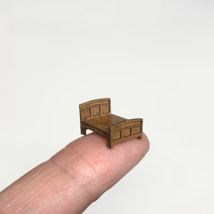 May include: A miniature wooden bed, crafted from brown cardboard, is displayed on a finger. The bed features a simple design with a headboard and footboard.