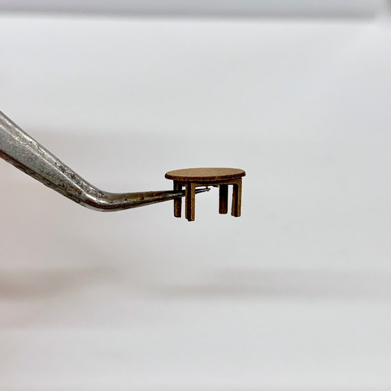 以下が含まれることがあります： 丸い天板と4本の脚を持つ、小さな茶色の木製テーブル。ピンセットで持ち上げられています。