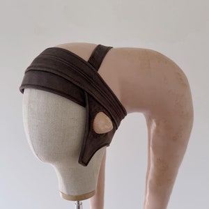 Puede incluir: Una diadema de tela marrón con una pieza de cabeza larga, curva y de color rosa claro. La diadema se lleva en una cabeza de maniquí.