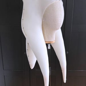 K&ouml;nnte beinhalten: Ein wei&szlig;es Stoffkost&uuml;mteil, m&ouml;glicherweise ein Kopfschmuck oder eine Kapuze, mit langen, spitzen Verl&auml;ngerungen. Die Kanten sind mit dunklen N&auml;hten umrandet. Der Artikel wird auf einem Schaufensterpuppenkopf und -st&auml;nder vor dunklem Hintergrund ausgestellt.