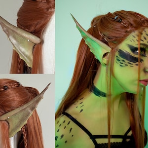 May include: A pair of green and gold elf ears, made of a flexible material, are shown on a model with red hair. The ears are pointed and have a textured surface.
