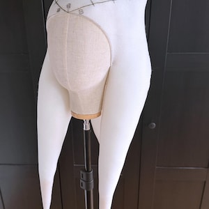 K&ouml;nnte beinhalten: Ein wei&szlig;es Stoff-Kopfst&uuml;ck mit langen, spitz zulaufenden Verl&auml;ngerungen, m&ouml;glicherweise ein Kost&uuml;mteil. Das Kopfst&uuml;ck ist auf einem Mannequinkopf montiert, der eine beige Stoffbasis freilegt. Das Design umfasst gestickte Details und ein dekoratives Element &uuml;ber der Stirn.