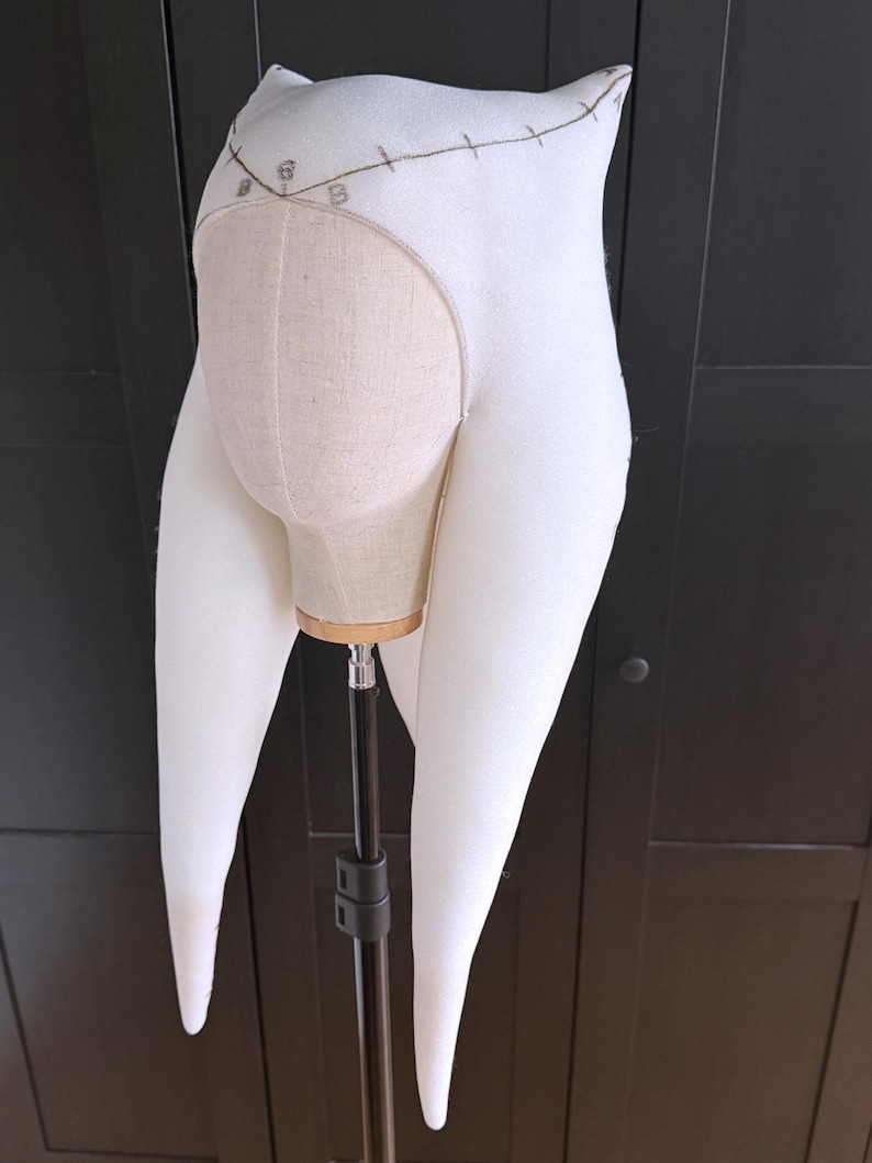 K&ouml;nnte beinhalten: Ein wei&szlig;es Stoff-Kopfst&uuml;ck mit langen, spitz zulaufenden Verl&auml;ngerungen, m&ouml;glicherweise ein Kost&uuml;mteil. Das Kopfst&uuml;ck ist auf einem Mannequinkopf montiert, der eine beige Stoffbasis freilegt. Das Design umfasst gestickte Details und ein dekoratives Element &uuml;ber der Stirn.