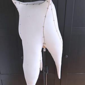 K&ouml;nnte beinhalten: Ein gro&szlig;er, wei&szlig;er Pl&uuml;schzahn mit schwarzen Nahtdetails. Der Zahn ist auf einem Metallst&auml;nder vor einem dunklen Hintergrund montiert. Die N&auml;hte umrei&szlig;en die Form des Zahns und heben seine Wurzeln und Krone hervor.