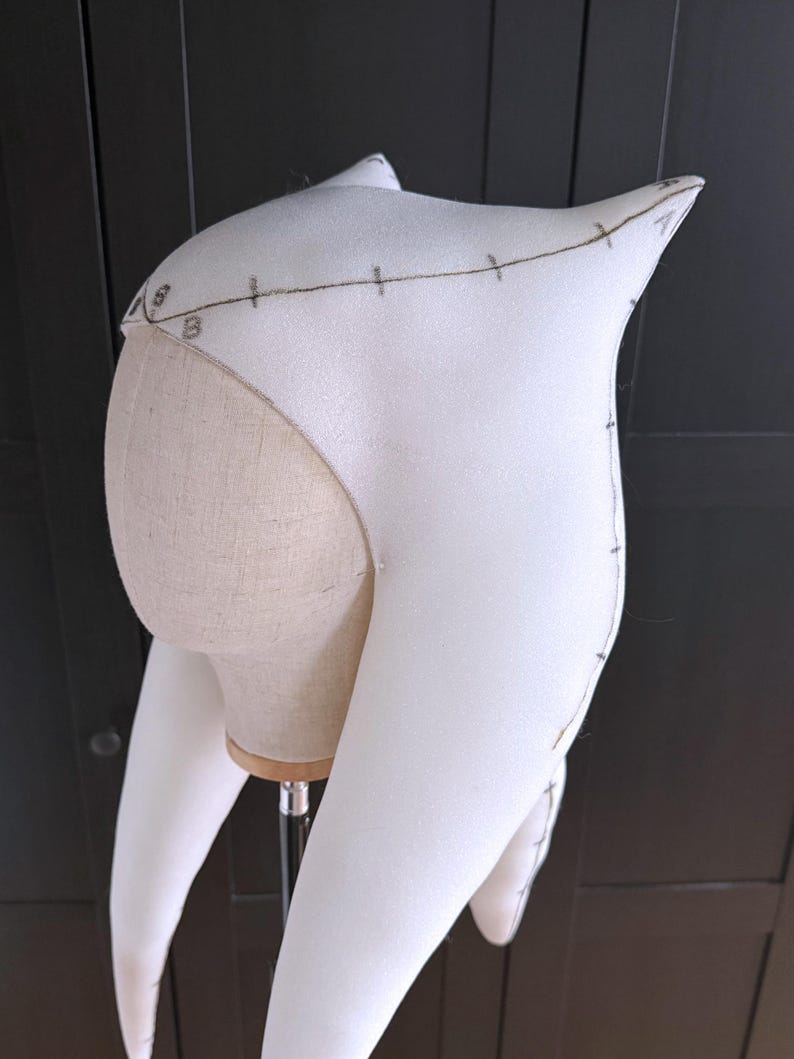 K&ouml;nnte beinhalten: Ein wei&szlig;es Schaumstoff-Kopfst&uuml;ck mit spitzen Ohren und langen, gebogenen Verl&auml;ngerungen. Das Kopfst&uuml;ck befindet sich auf einem Schaufensterpuppenkopf. Braune Linien und Zahlen sind auf den Schaumstoff gezeichnet und weisen auf Ma&szlig;e und Designdetails hin. Der Hintergrund ist dunkles Holz.