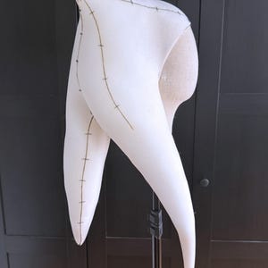 K&ouml;nnte beinhalten: Ein wei&szlig;es, hornf&ouml;rmiges Kost&uuml;mteil mit dunklen Nahtdetails. Der Stoff ist &uuml;ber eine Form gespannt, die die Form und Konstruktion zeigt. Das Design weist lange, gebogene H&ouml;rner auf, die auf ein Fantasy- oder Theater-Thema hindeuten.