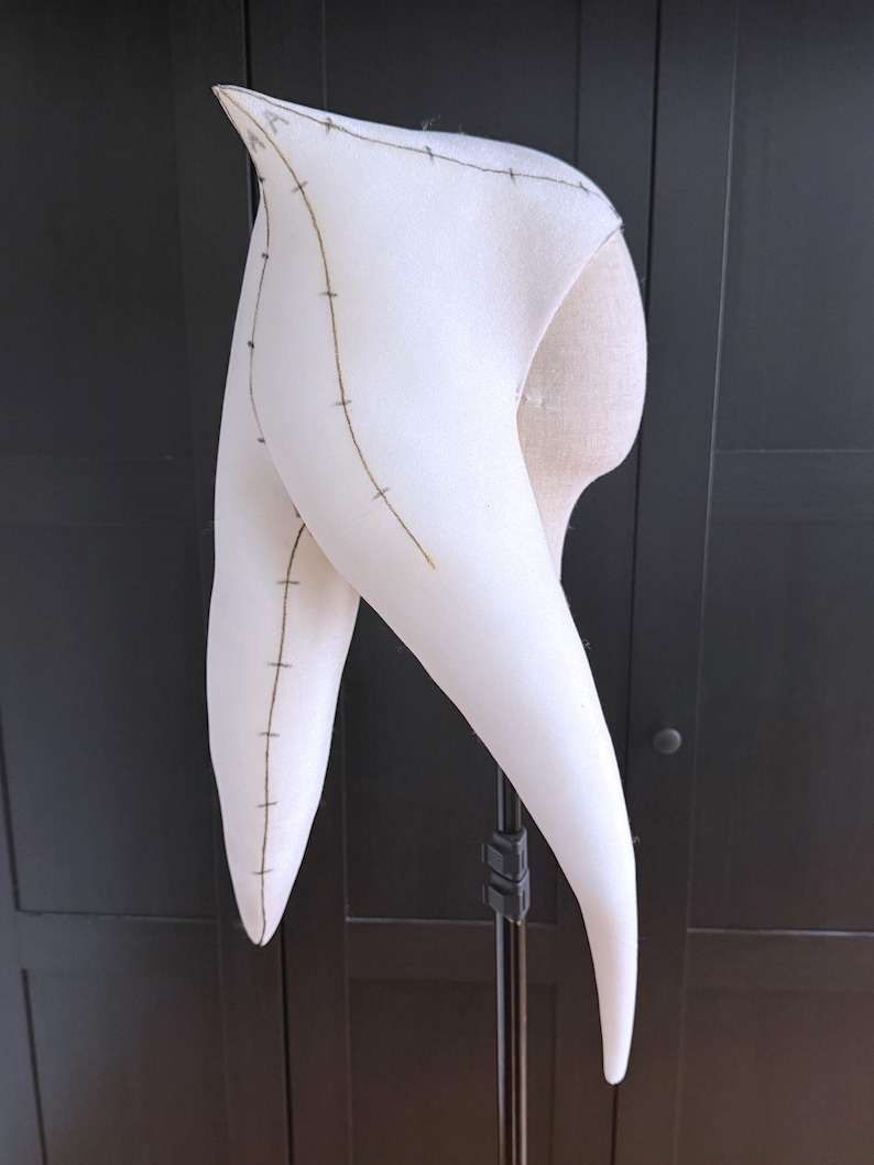 K&ouml;nnte beinhalten: Ein wei&szlig;es, hornf&ouml;rmiges Kost&uuml;mteil mit dunklen Nahtdetails. Der Stoff ist &uuml;ber eine Form gespannt, die die Form und Konstruktion zeigt. Das Design weist lange, gebogene H&ouml;rner auf, die auf ein Fantasy- oder Theater-Thema hindeuten.