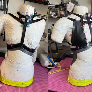 May include: A black leather harness with straps and clips, pinned to a white dress form. The harness is being fitted for sewing.