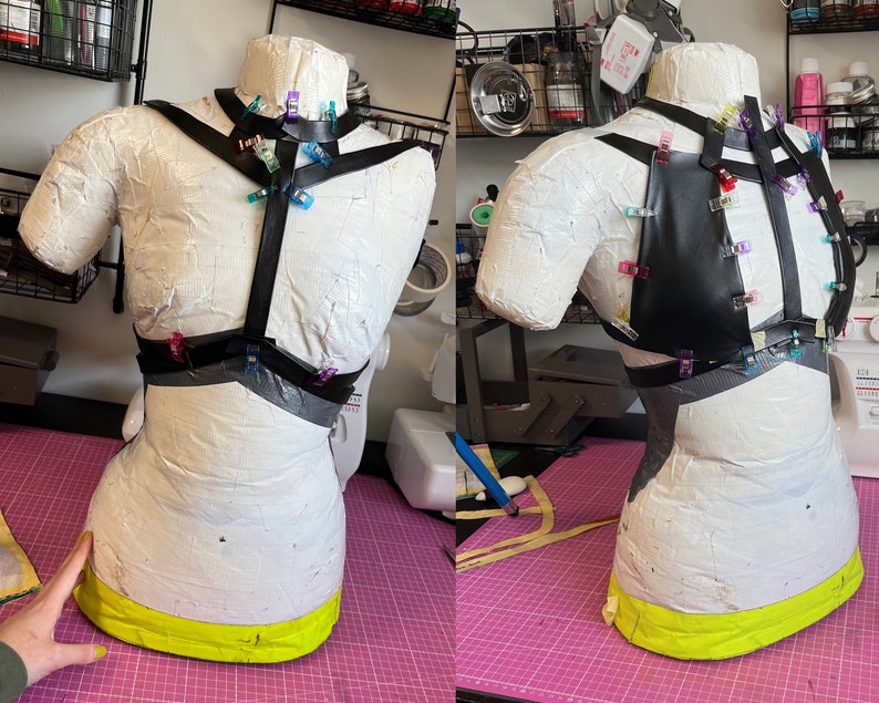 May include: A black leather harness with straps and clips, pinned to a white dress form. The harness is being fitted for sewing.